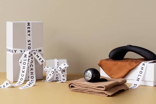 Boîtes cadeaux avec rubans 'Stockholm', un appareil noir tenu à la main, une écharpe orange et un tissu plié marron sur un fond beige.