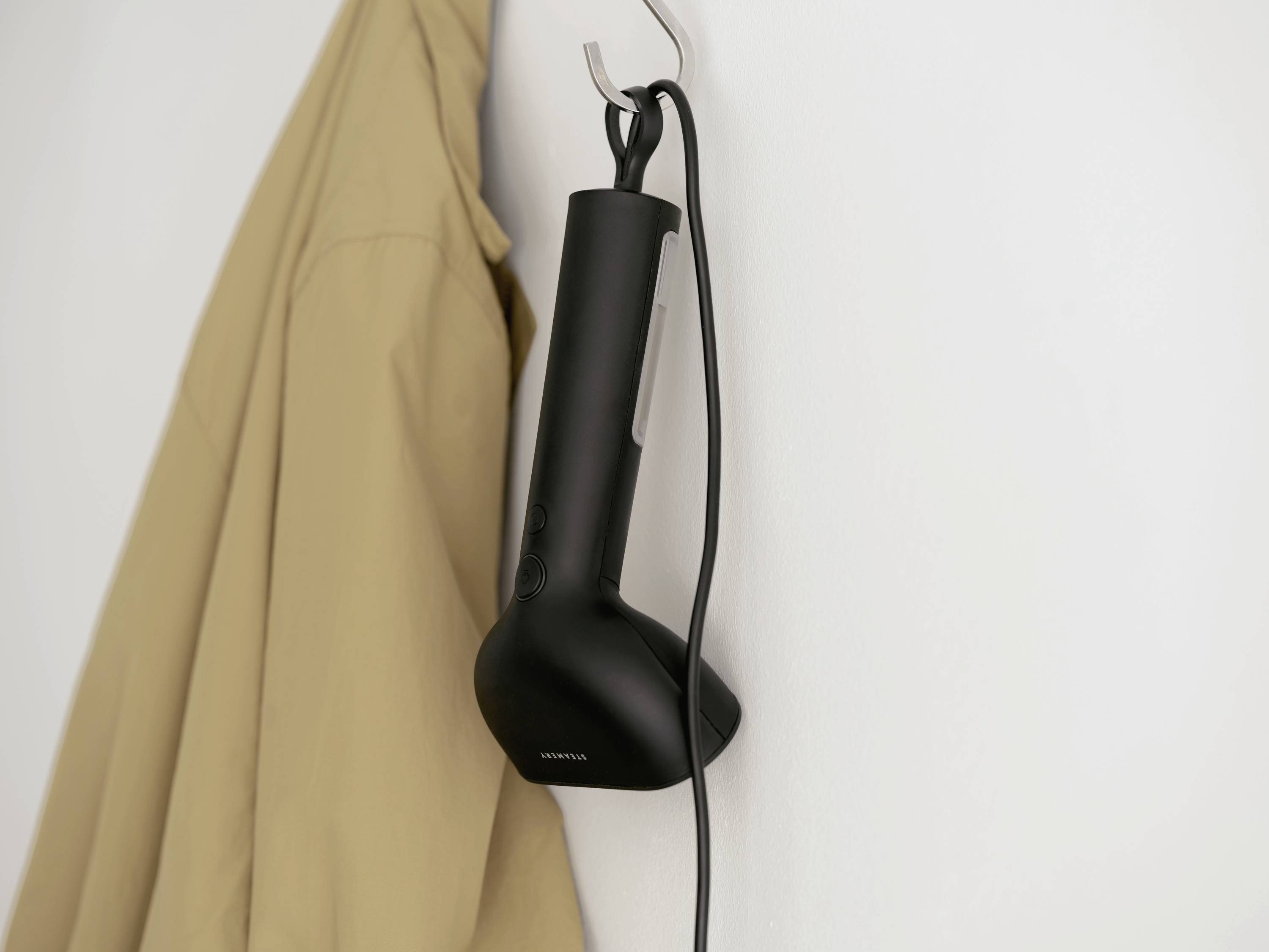 Un fer à repasser vapeur noir est suspendu à un crochet sur le mur. À côté, un manteau beige est accroché.