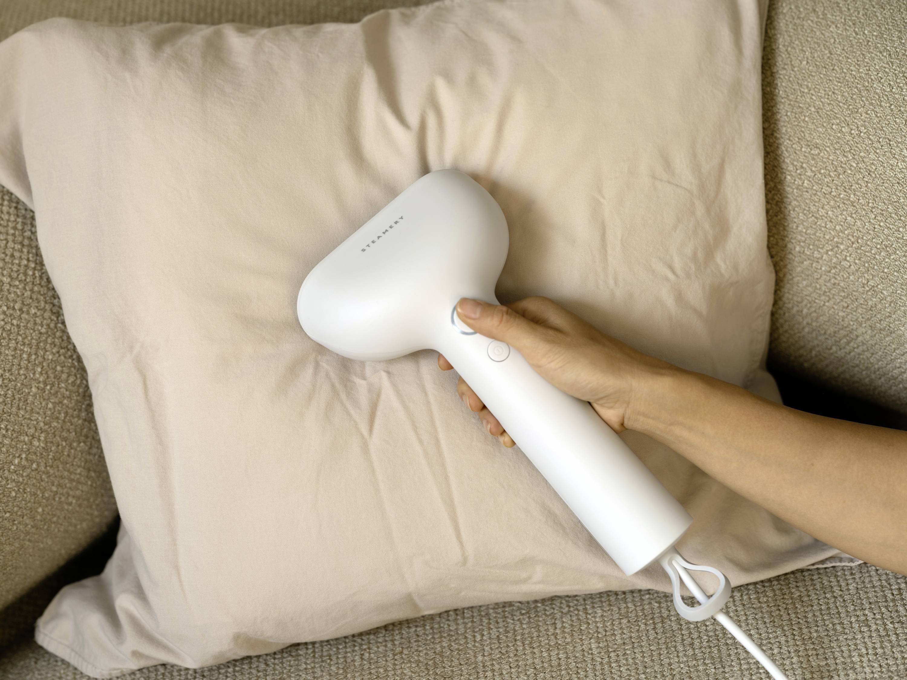 Une main tient un fer à repasser à vapeur blanc contre un coussin sur un canapé beige.
