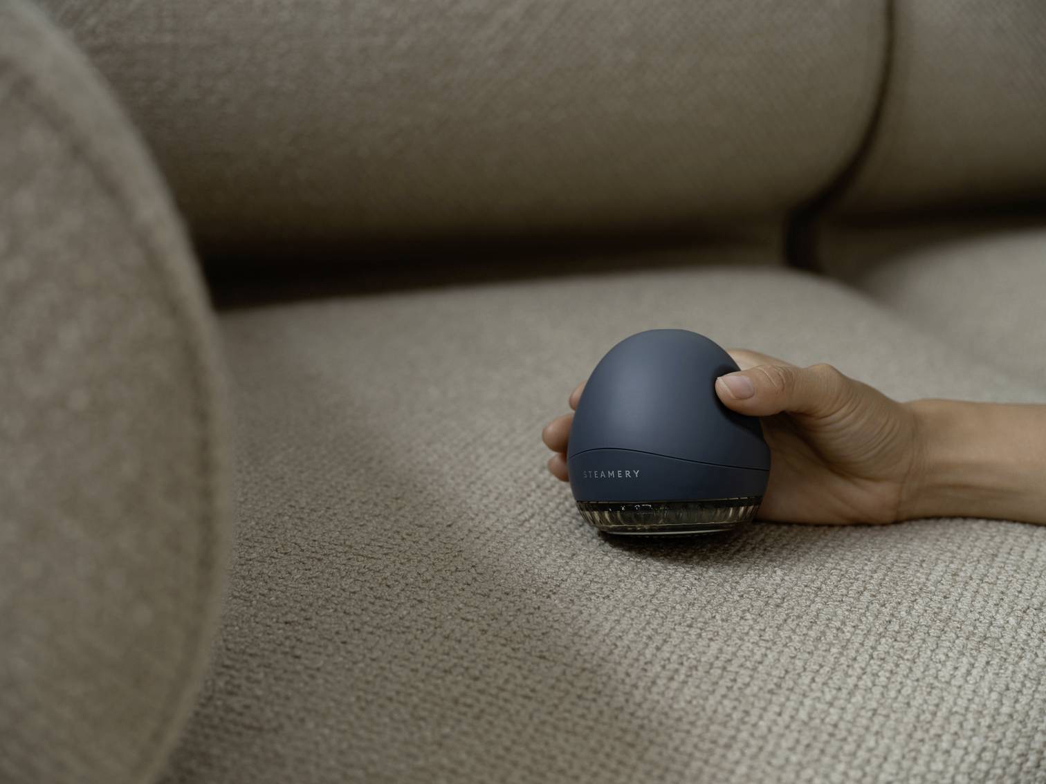 Une main tient une petite machine ronde bleue sur un canapé beige. Elle semble être utilisée pour nettoyer le tissu.