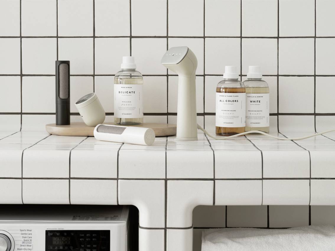 Une salle de bain avec un mur carrelé présente des produits de soin et des accessoires sur une étagère. Au premier plan se trouve un appareil à main.