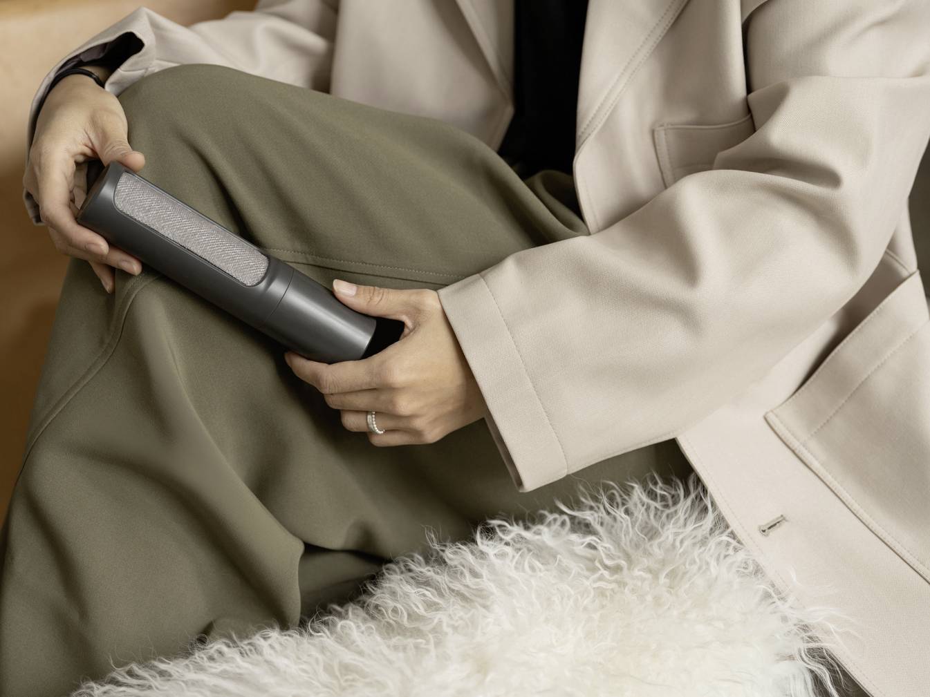 Une personne tient un haut-parleur portable sans fil dans sa main, assise sur un canapé, portant un manteau beige et un pantalon vert.