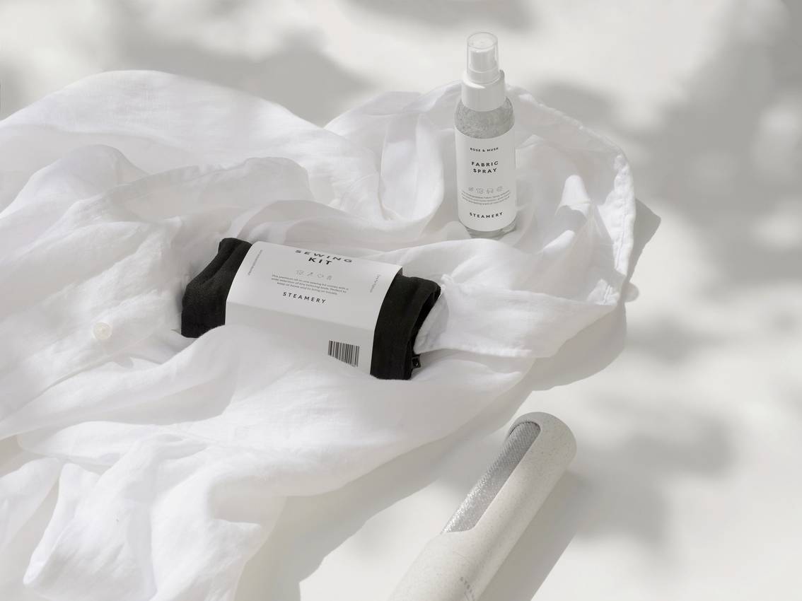 Une chemise blanche est posée à plat, à côté d'un vaporisateur et d'un produit de soin emballé. Des jeux d'ombres créent une ambiance douce.