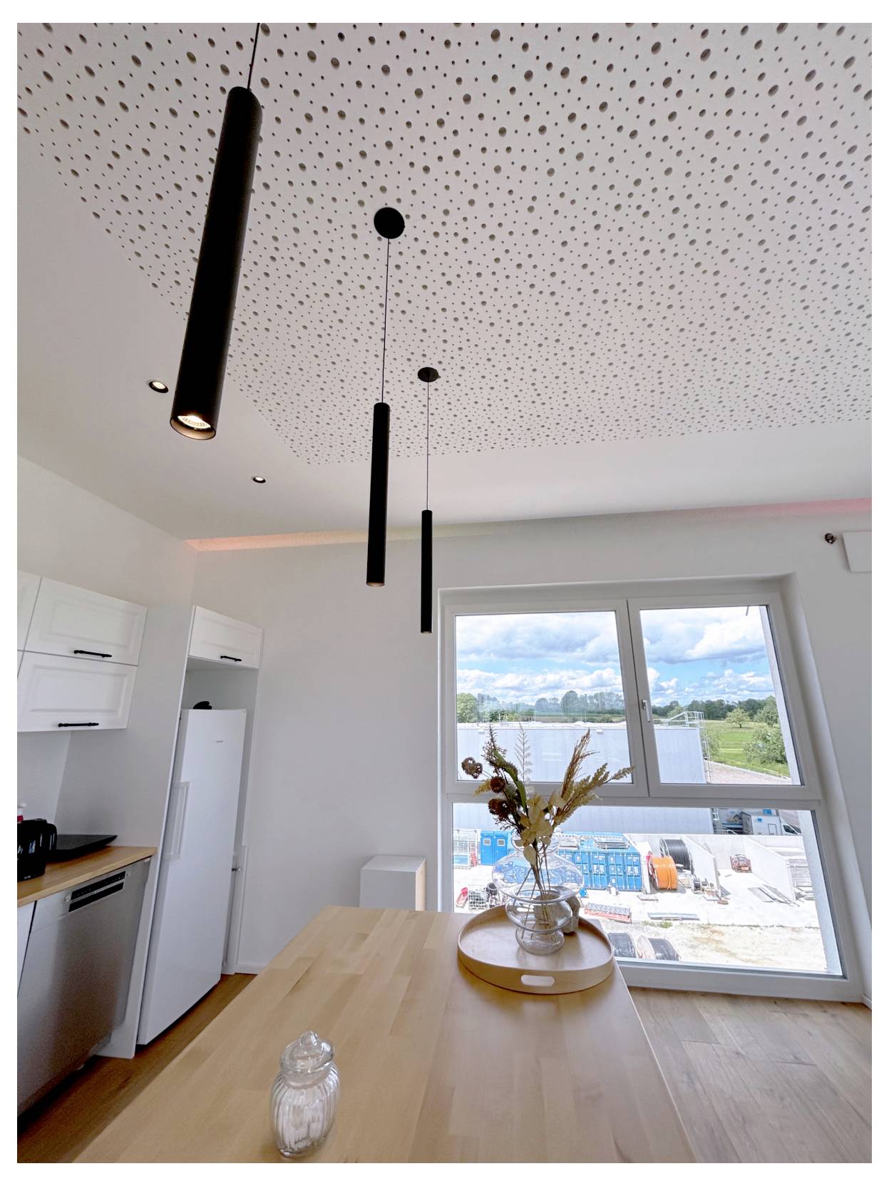 Cuisine moderne avec îlot en bois et suspensions, donnant sur une fenêtre dont la vue embrasse un chantier à l'extérieur.