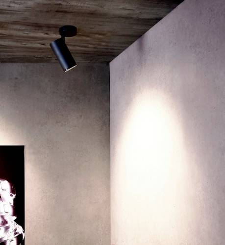 Un luminaire noir au plafond éclaire un mur rose dans un espace moderne. À gauche, une partie d'un objet brillant est visible.