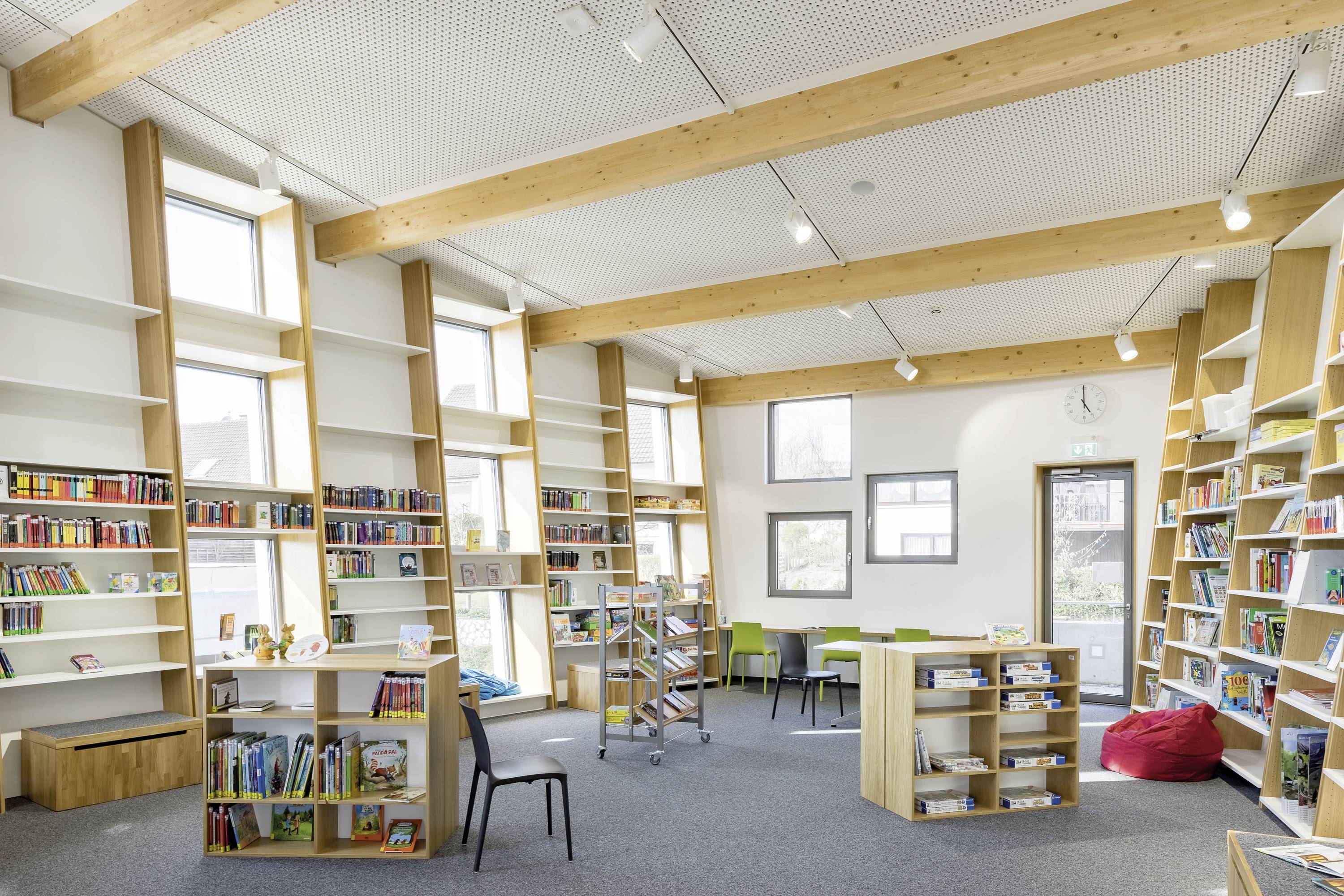 Espace bibliothécaire lumineux et moderne avec des étagères remplies de livres, des tables et des chaises pour la lecture. De grandes fenêtres laissent entrer beaucoup de lumière naturelle.