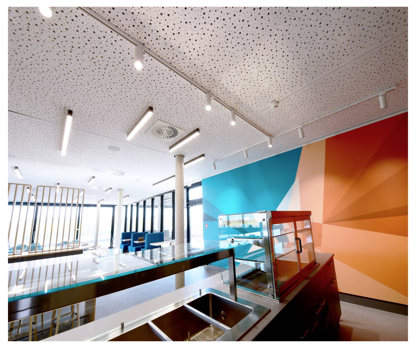 Intérieur moderne de cafétéria avec comptoir de service, éviers en acier inoxydable, sièges bleus et fresque murale géométrique lumineuse aux tons turquoise et orange.