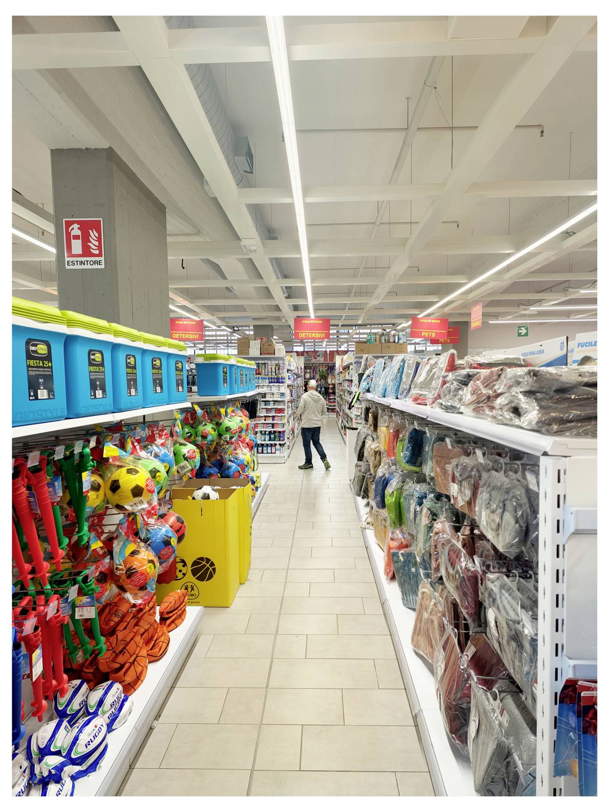 Une allée de magasin de jouets avec des étagères de balles et d'accessoires colorés à gauche et des articles emballés à droite, un client s'éloignant au centre.
