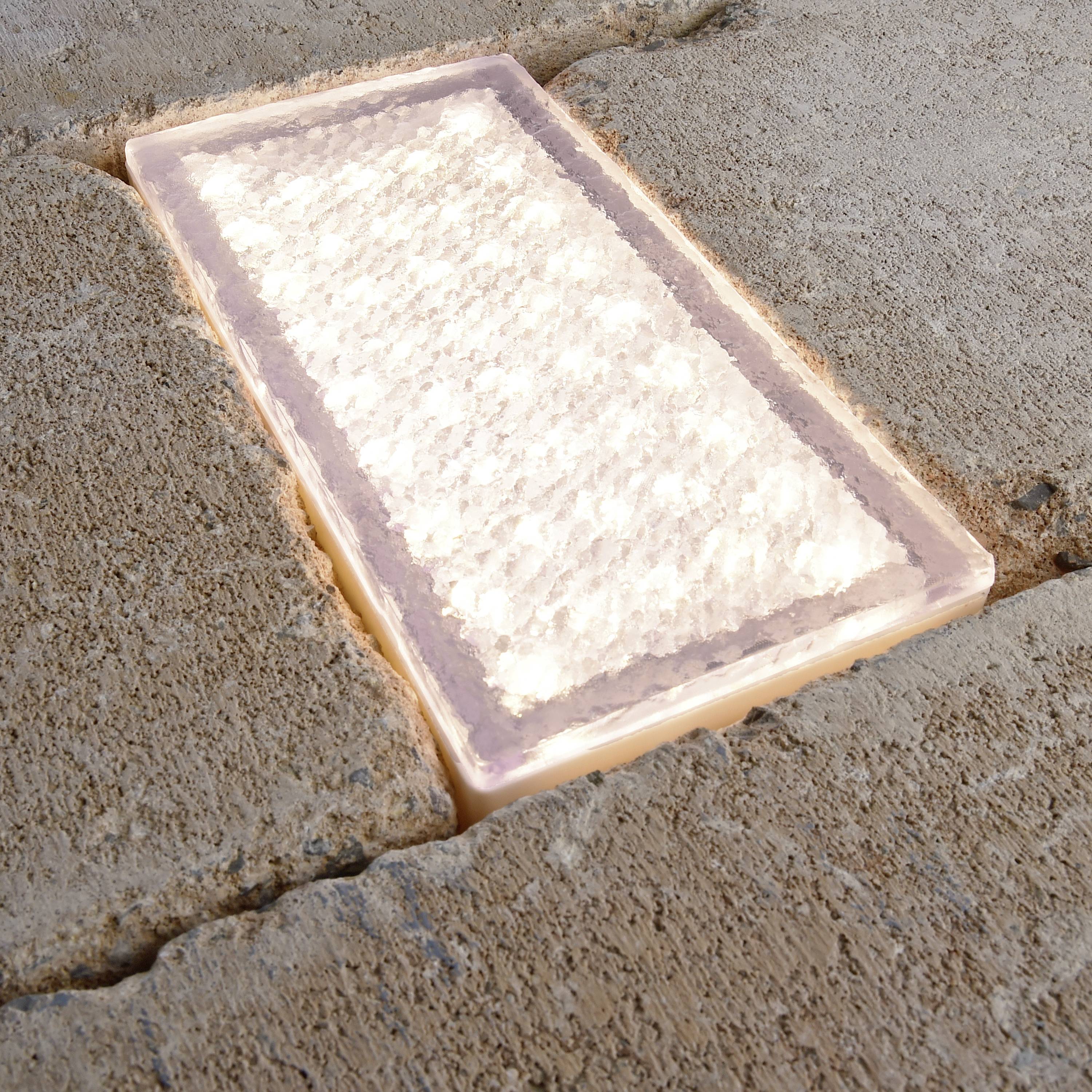 Une pierre de pavage rectangulaire illuminée, encastrée dans un trottoir composé de pierres aux formes irrégulières, s'illumine au crépuscule.