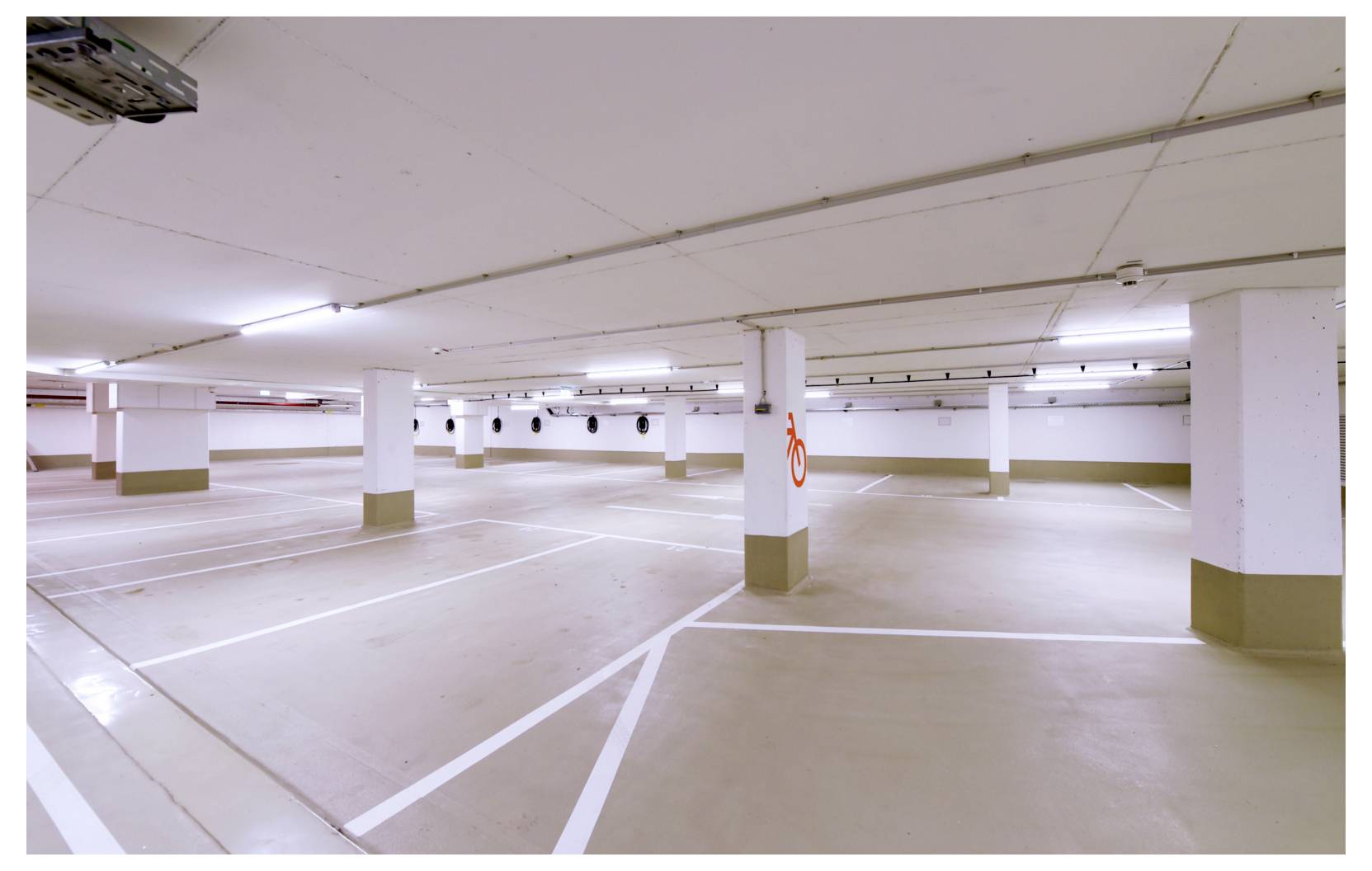 Parking souterrain vide avec des places de stationnement peintes, des piliers blancs et des plafonniers fluorescents.
