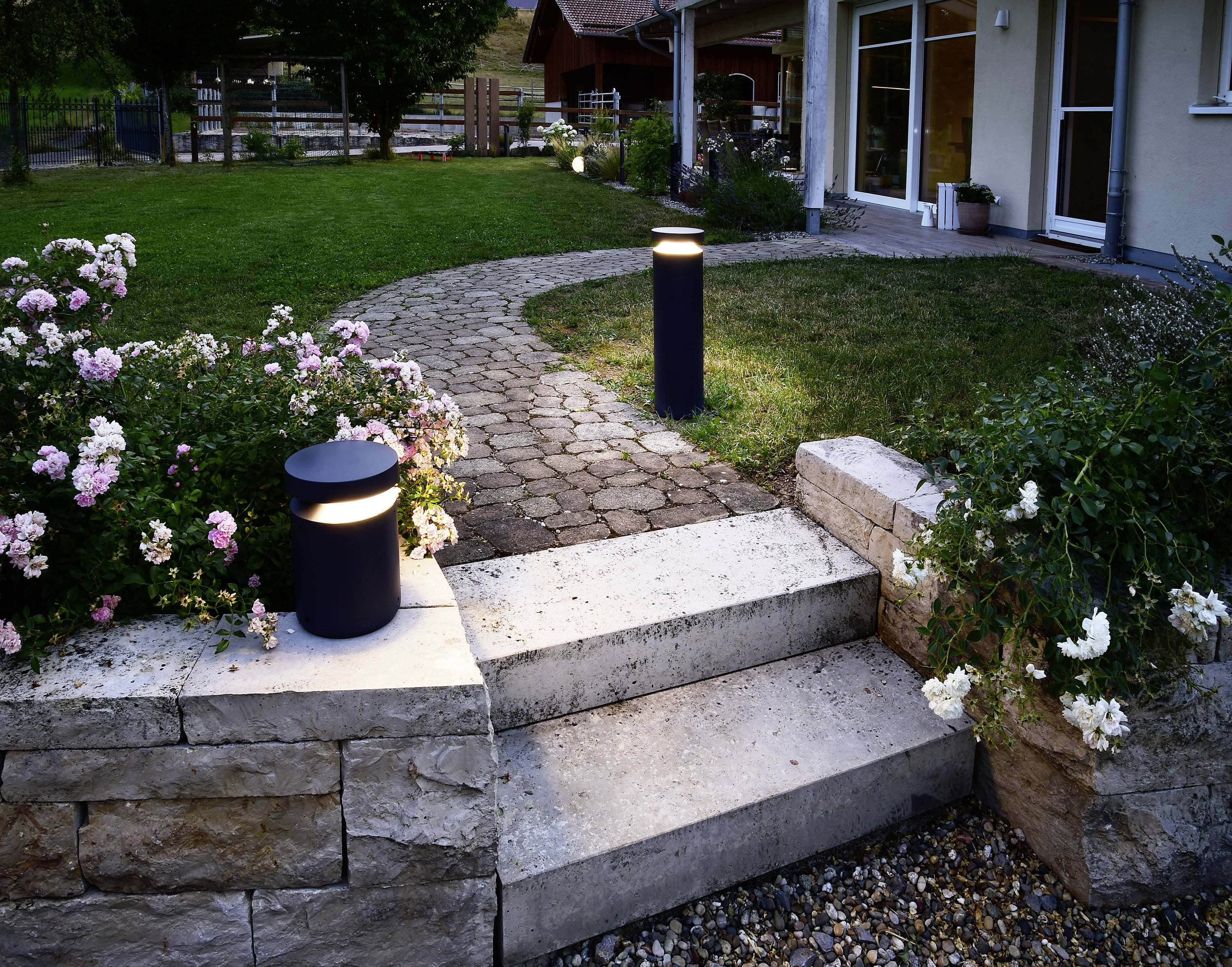 Un chemin de jardin en dalles de pierre mène à une maison. Deux lampes extérieures lumineuses éclairent le chemin, entourées de parterres de fleurs.