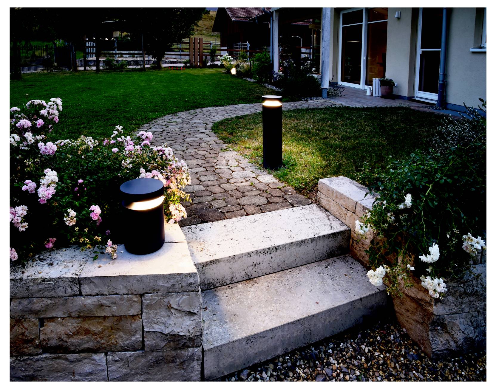 Allée de jardin avec bornes lumineuses le long du chemin, marches en pierre et parterres de fleurs menant à une maison au crépuscule.