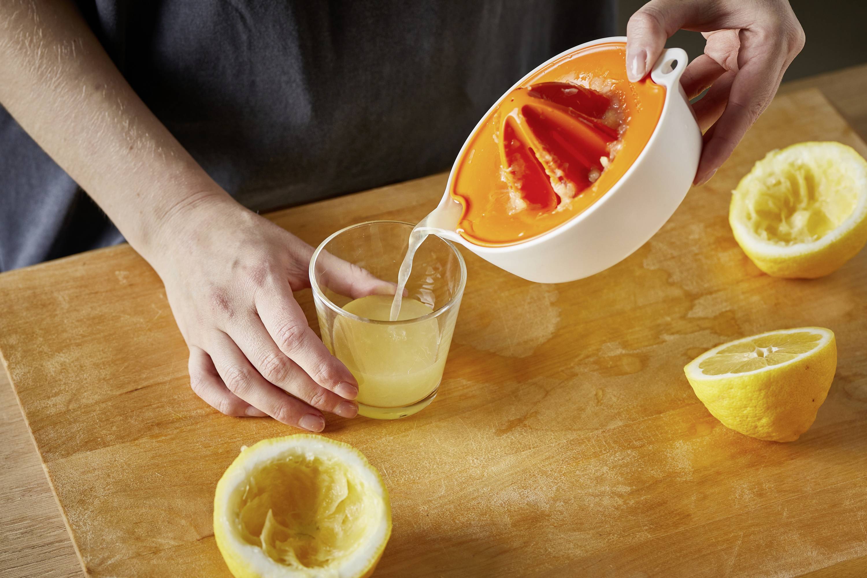 Une personne presse des citrons avec un presse-agrumes manuel. Le jus est versé dans un verre. Deux moitiés de citrons évidées sont visibles.