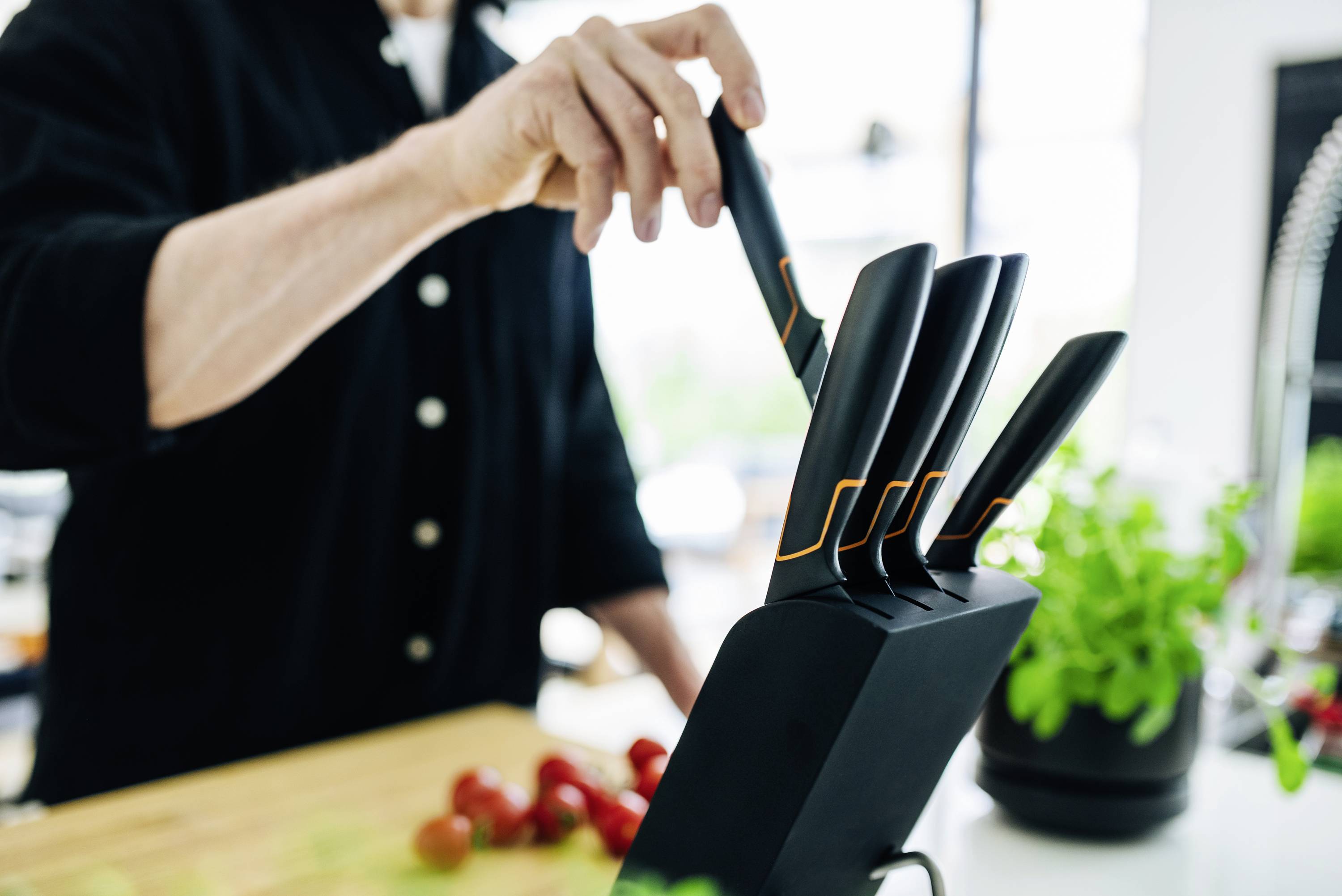 Une personne habillée en noir saisit un couteau dans un bloc à couteaux dans une cuisine moderne. Des tomates et des plantes aromatiques sont visibles en arrière-plan.