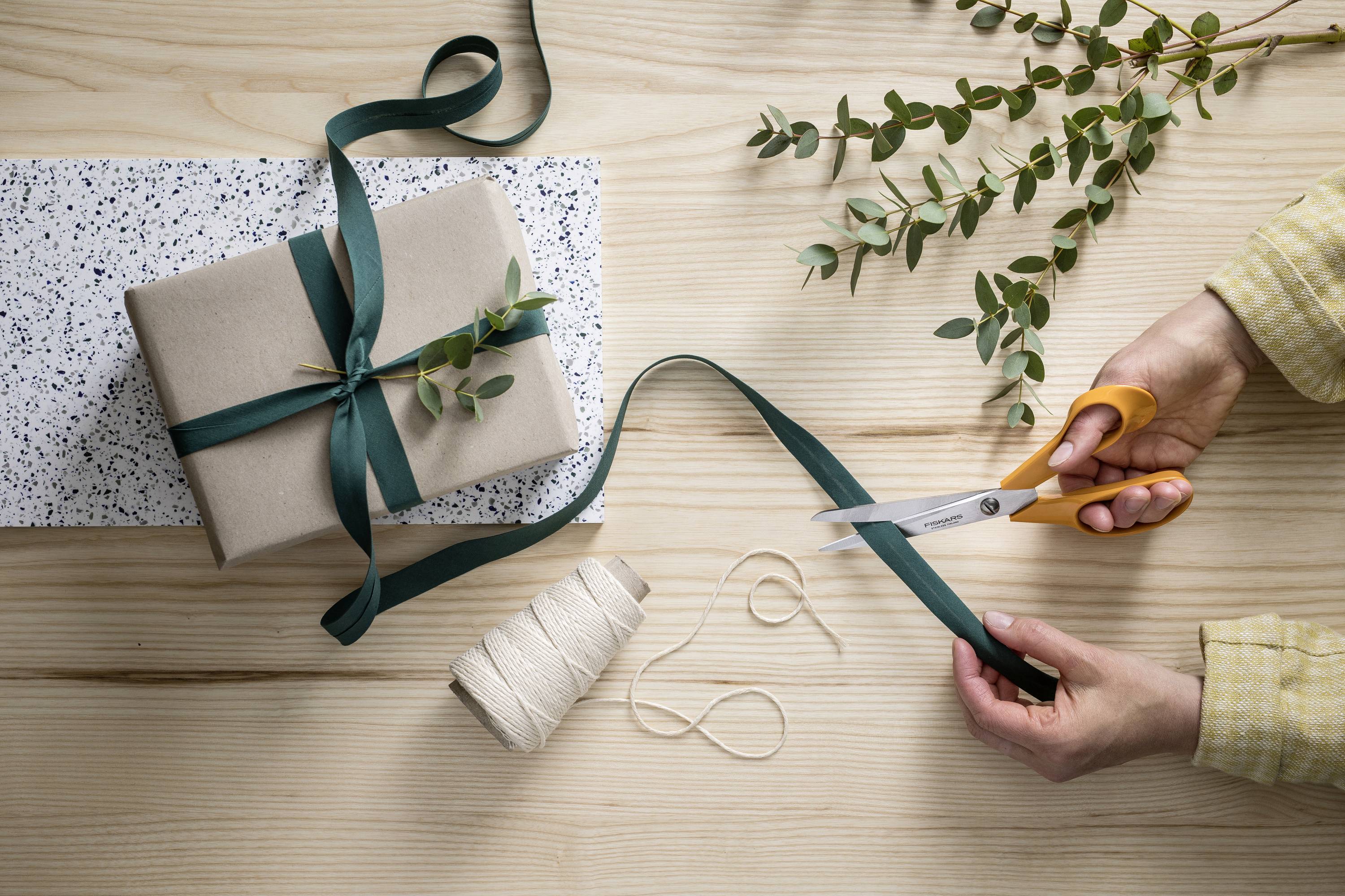 Une personne coupe un ruban vert avec des ciseaux. Un cadeau emballé est posé sur un papier à motifs, décoré de branches d'eucalyptus.