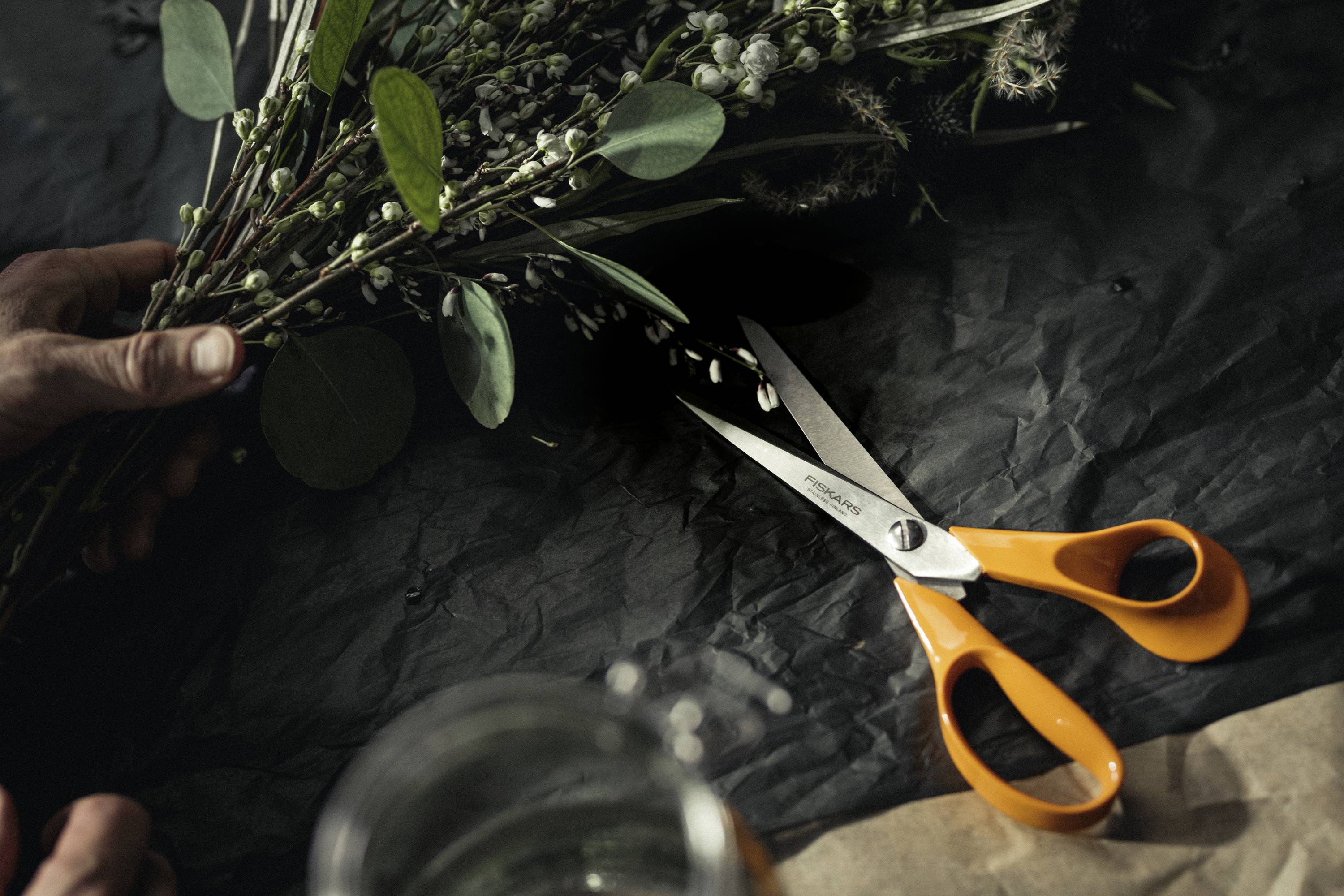Une personne tient un bouquet de fleurs. À côté, des ciseaux orange reposent sur un fond sombre.
