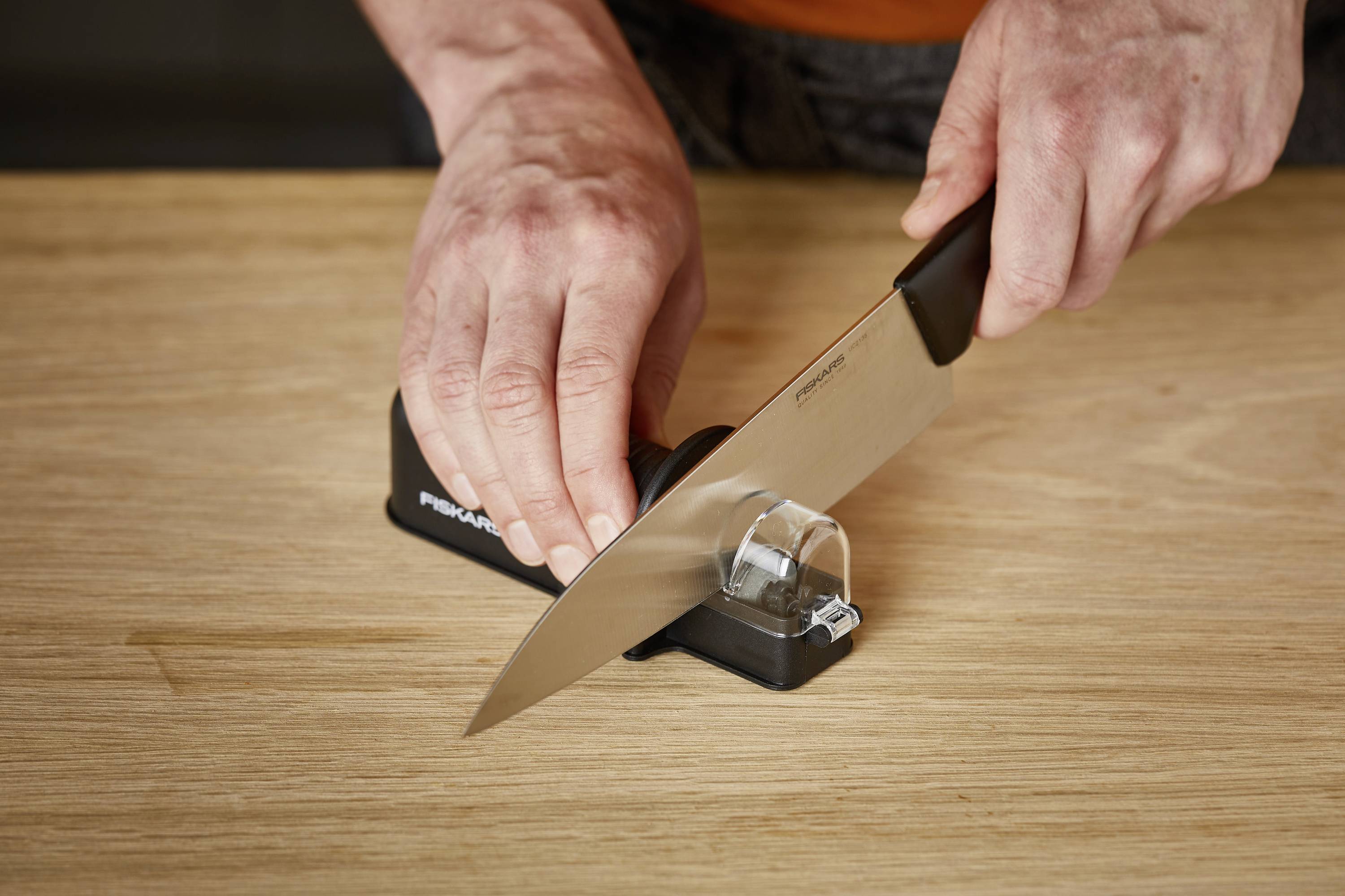 Une personne aiguise un couteau de cuisine avec un aiguiseur manuel sur une table en bois.