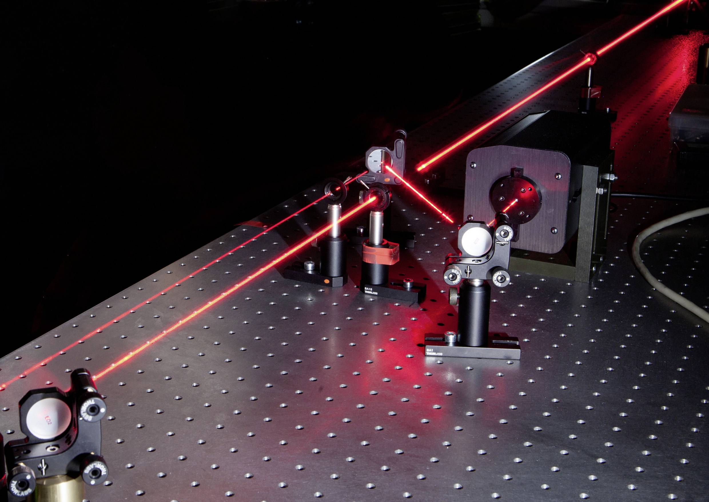 Un montage optique de laboratoire montre des faisceaux laser rouges dirigés à travers des lentilles et des miroirs, pour étudier le comportement de la lumière.