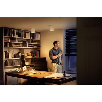 Un homme se tient près d'une fenêtre, tenant une boisson à la main, dans un bureau dominé par un grand bureau et des étagères de livres.
