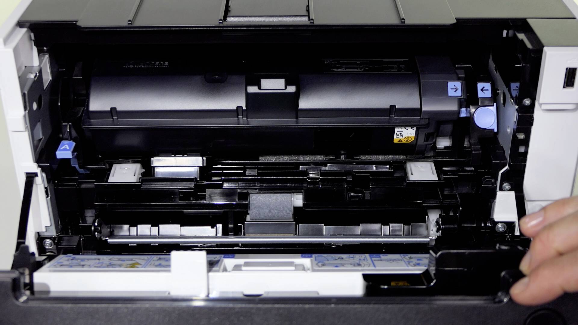 Une imprimante ouverte montrant ses composants internes tels que la cartouche de toner et le système d'alimentation papier. Une main maintient le capot ouvert.