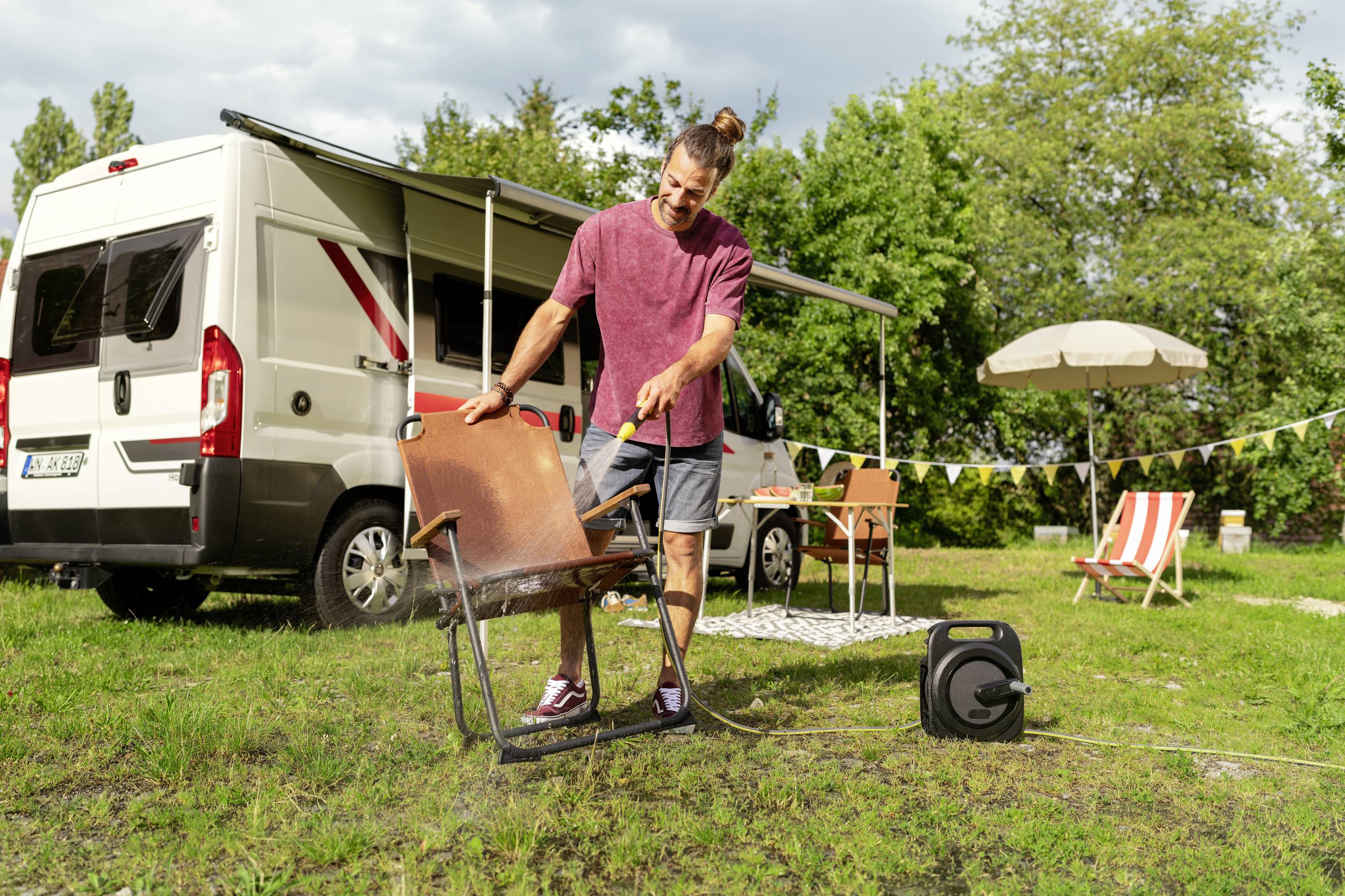 Un homme nettoie une chaise en bois avec un tuyau d'arrosage sur une pelouse à côté d'un camping-car, avec des arbres et un parasol en arrière-plan.