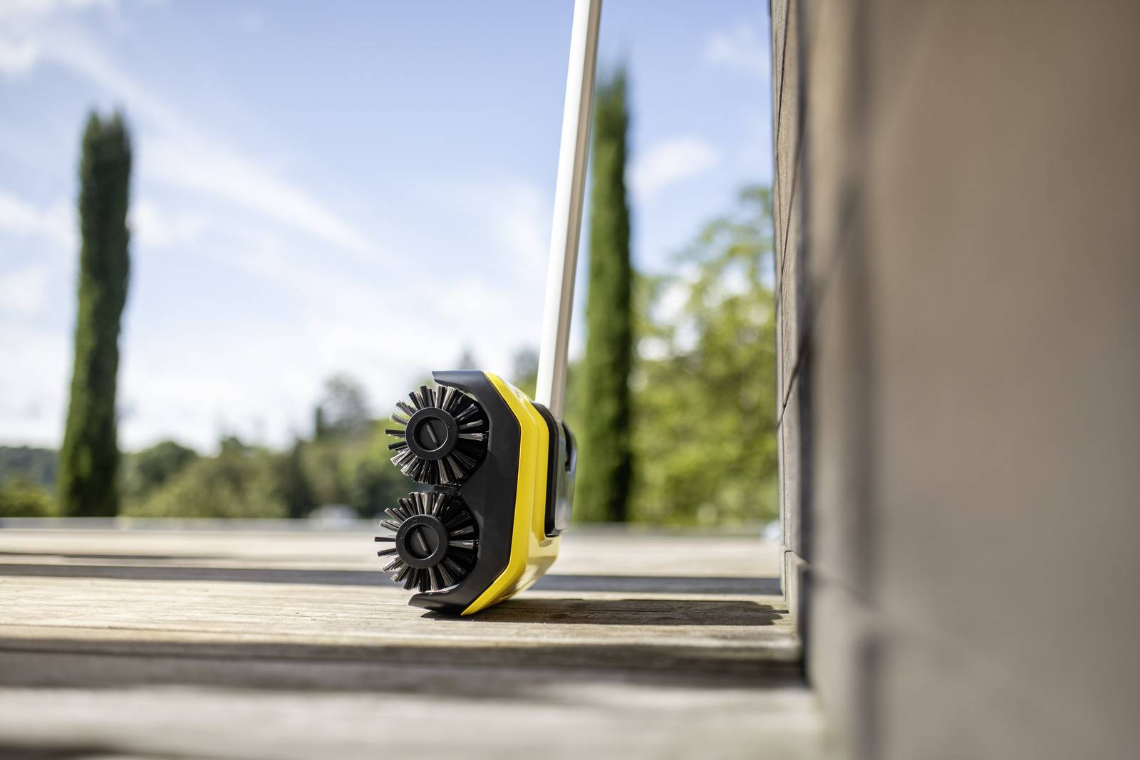 Un balai-brosse jaune avec deux brosses rondes est poussé sur une terrasse en bois, avec des arbres visibles en arrière-plan.