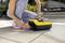 Une personne nettoie le sol d'une terrasse avec une machine de nettoyage jaune. Des plantes et des meubles de jardin sont visibles en arrière-plan.