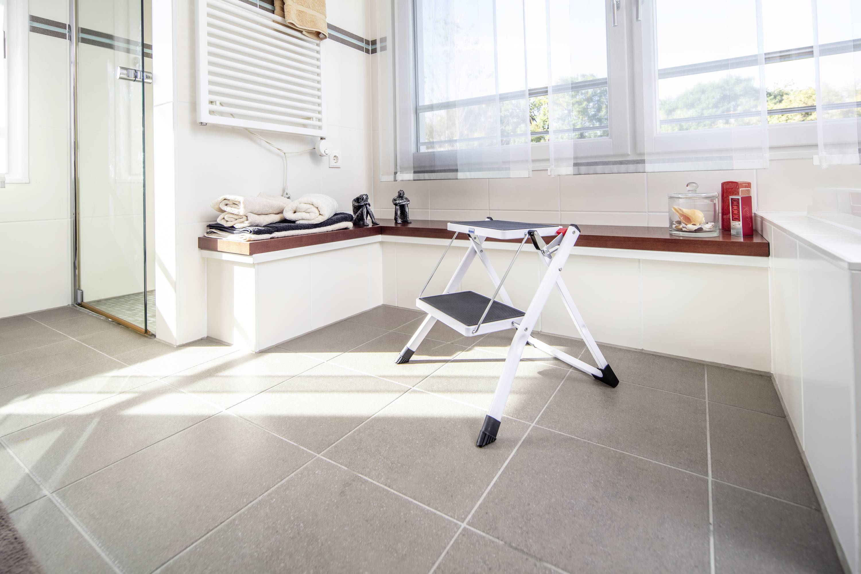 Salle de bain claire avec un tabouret au centre, serviettes sur le banc, douche à gauche, fenêtre en arrière-plan.