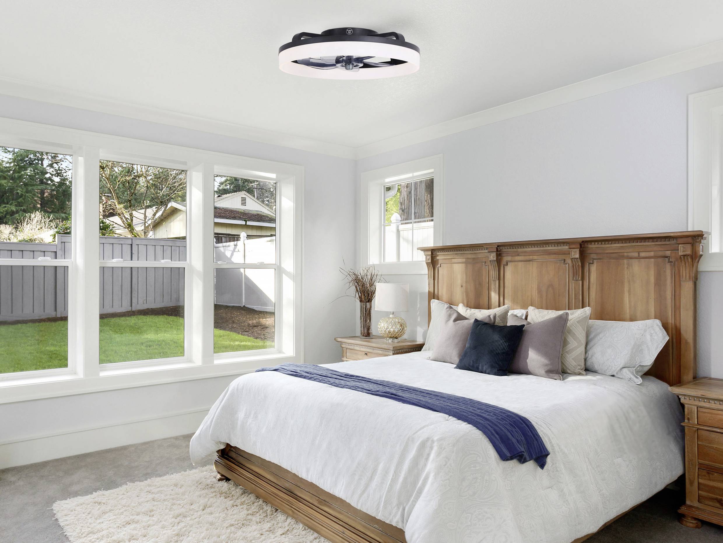 Chambre à coucher avec une grande fenêtre, un lit en bois et un ventilateur de plafond moderne. La pièce dégage une atmosphère calme et lumineuse.