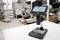 Microscope numérique sur une table dans un laboratoire technique, avec une carte de circuit imprimé sous l'objectif. À l'arrière-plan, différentes stations de travail sont visibles.