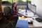 Une femme est assise dans un bureau devant un bureau, examinant des documents sur deux écrans. À l'arrière-plan, une fenêtre donne sur l'extérieur.