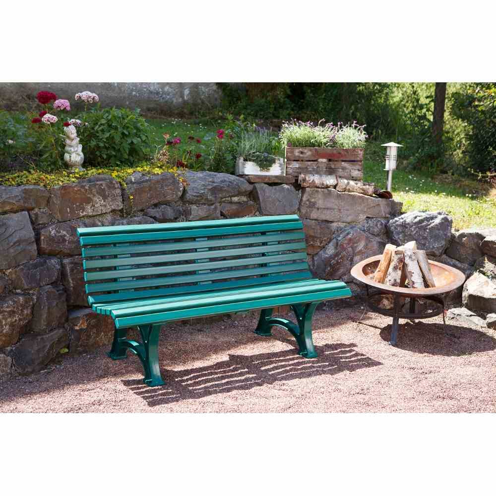 Un banc de jardin vert est placé devant un muret de pierre bas, décoré de fleurs. À côté se trouve un petit foyer circulaire.