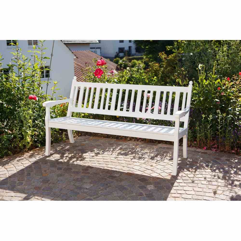 Un banc de jardin blanc est placé sur une place pavée, entouré de plantes hautes et d'une maison bleue en arrière-plan.