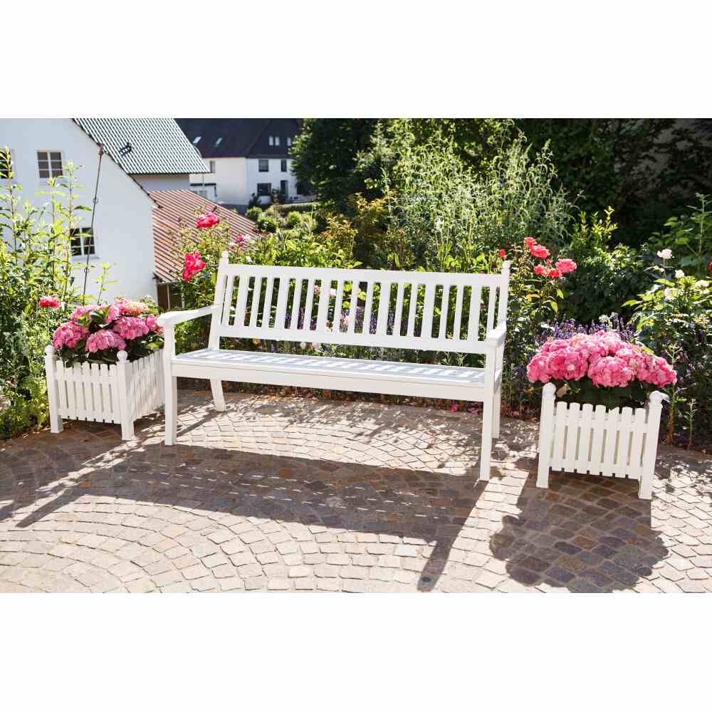 Banc de jardin blanc en bois sur des pavés, flanqué de deux jardinières contenant des fleurs roses. L'arrière-plan montre des plantes vertes et des maisons.