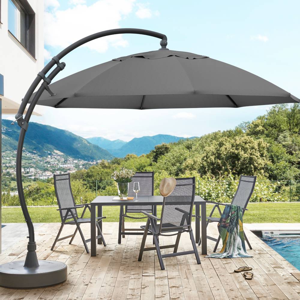 Table de jardin ronde avec quatre chaises et un grand parasol sur une terrasse. En arrière-plan, des montagnes et des arbres, un ciel bleu.