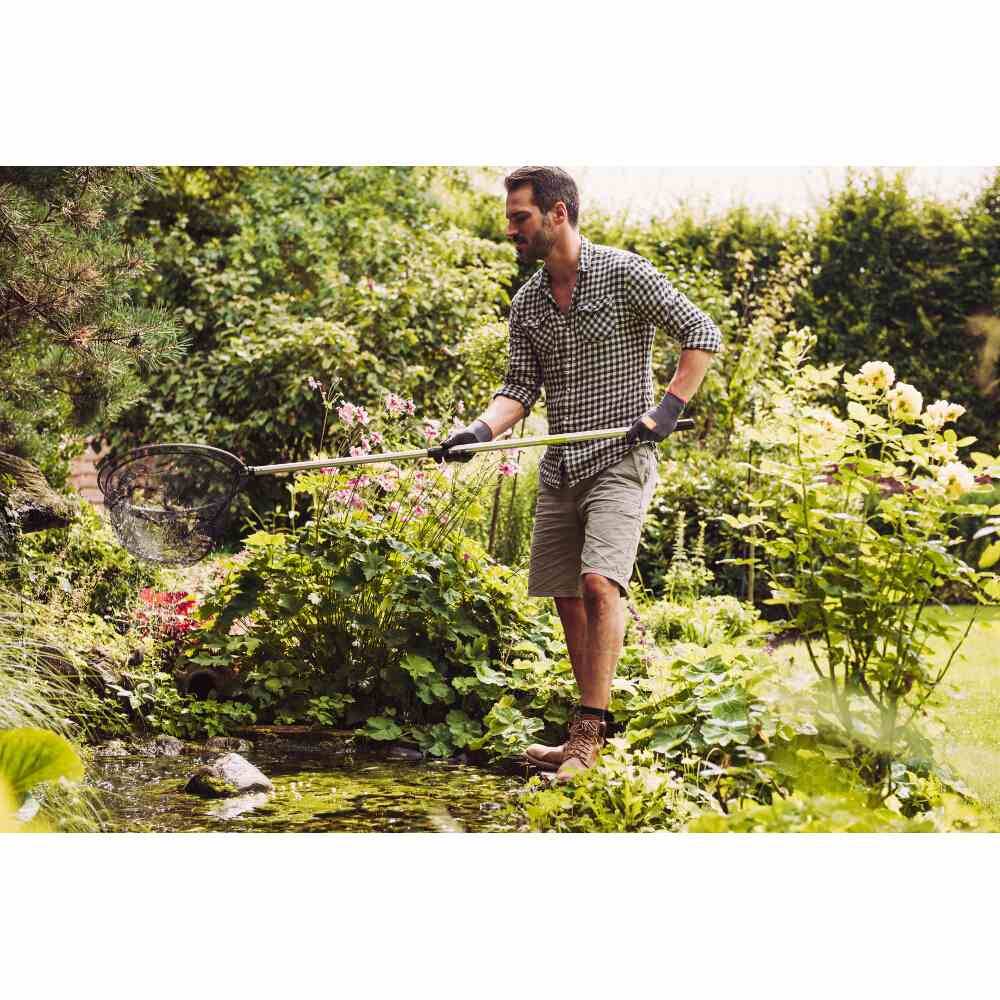 Un homme nettoie un étang avec une épuisette dans un jardin. Entouré de verdure luxuriante et de plantes, il porte des gants et des vêtements décontractés.