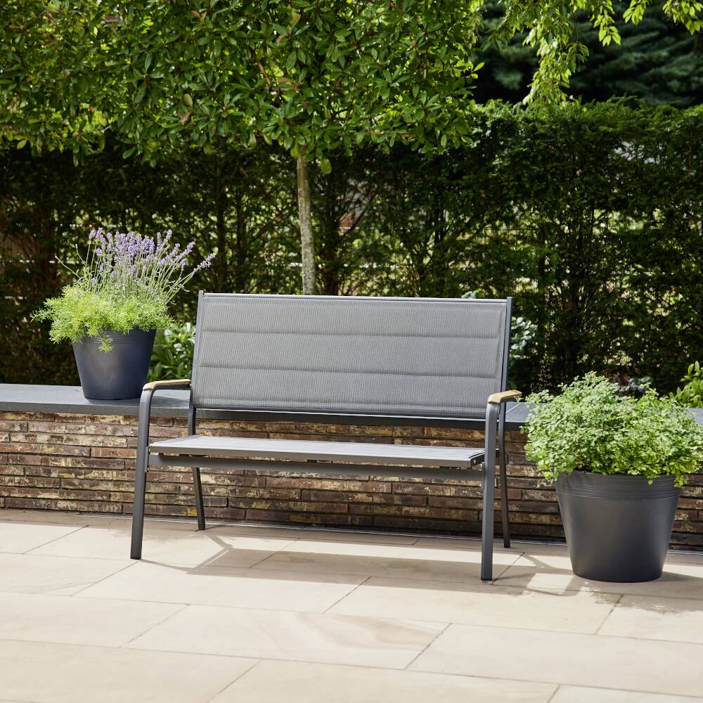 Un banc de jardin gris en métal est installé sur une terrasse. À côté se trouvent deux grands pots de fleurs contenant de la lavande et d'autres plantes. En arrière-plan, on peut voir une haie verte.