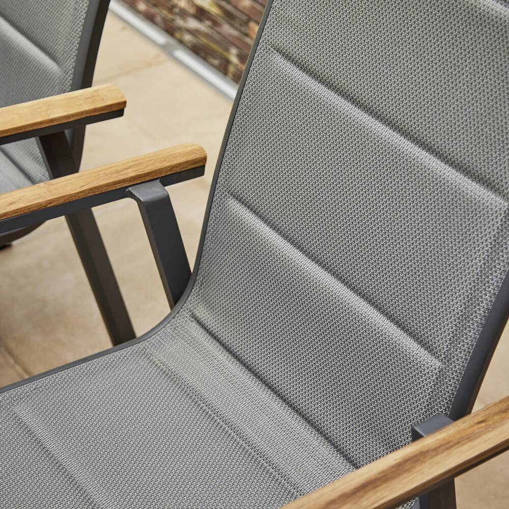 Une chaise de jardin grise avec des accoudoirs en bois est placée sur une terrasse. L'assise et le dossier sont réalisés en un matériau textile matelassé.