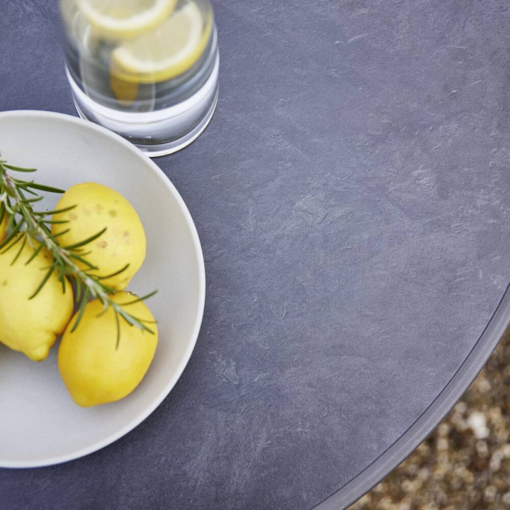 Un verre d'eau avec des tranches de citron et une assiette contenant trois citrons et une branche de romarin sur une table grise.