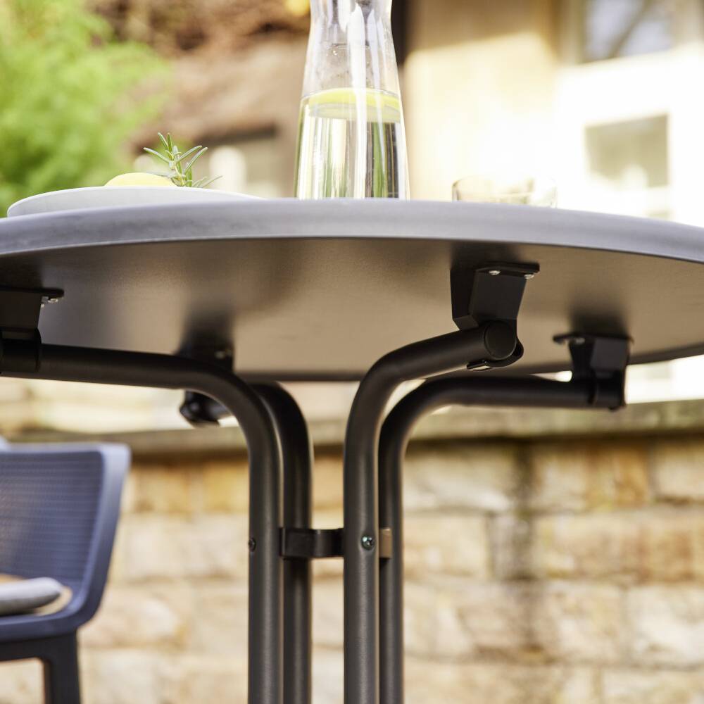 Une table ronde avec une bouteille d'eau en verre et deux verres posés dessus, à l'extérieur sur une terrasse avec un arrière-plan flou et une chaise.