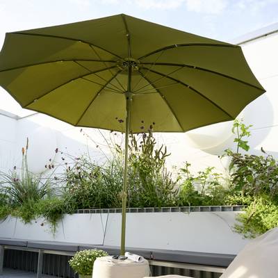 Un grand parasol vert est installé sur une terrasse, entouré de plantes. C'est une journée ensoleillée avec quelques nuages dans le ciel.