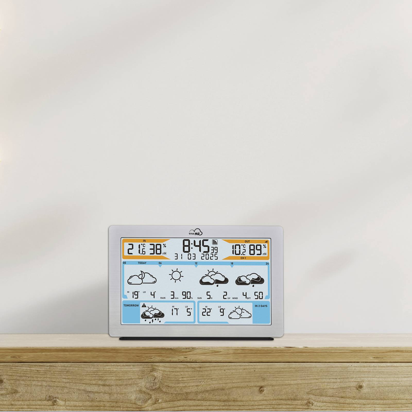 Une station météorologique numérique posée sur une table en bois affiche la température actuelle de 21,6°C et une prévision avec des symboles de nuages.