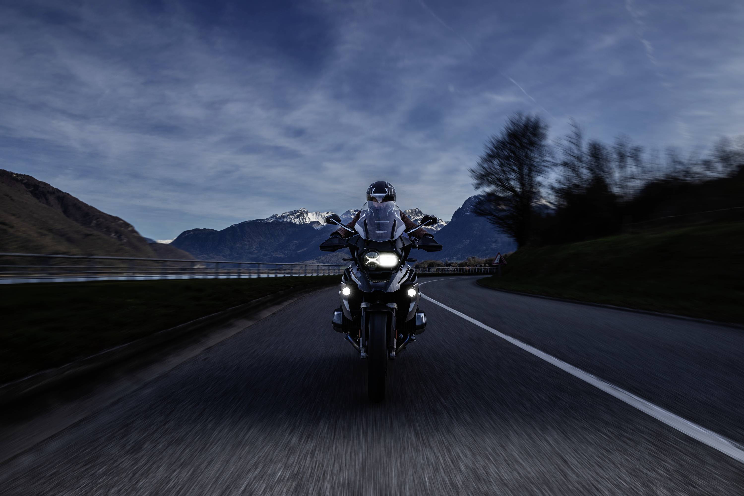 Motocycliste sur une route de montagne au crépuscule, phares allumés, roulant en direction du spectateur. Montagnes et arbres en arrière-plan.