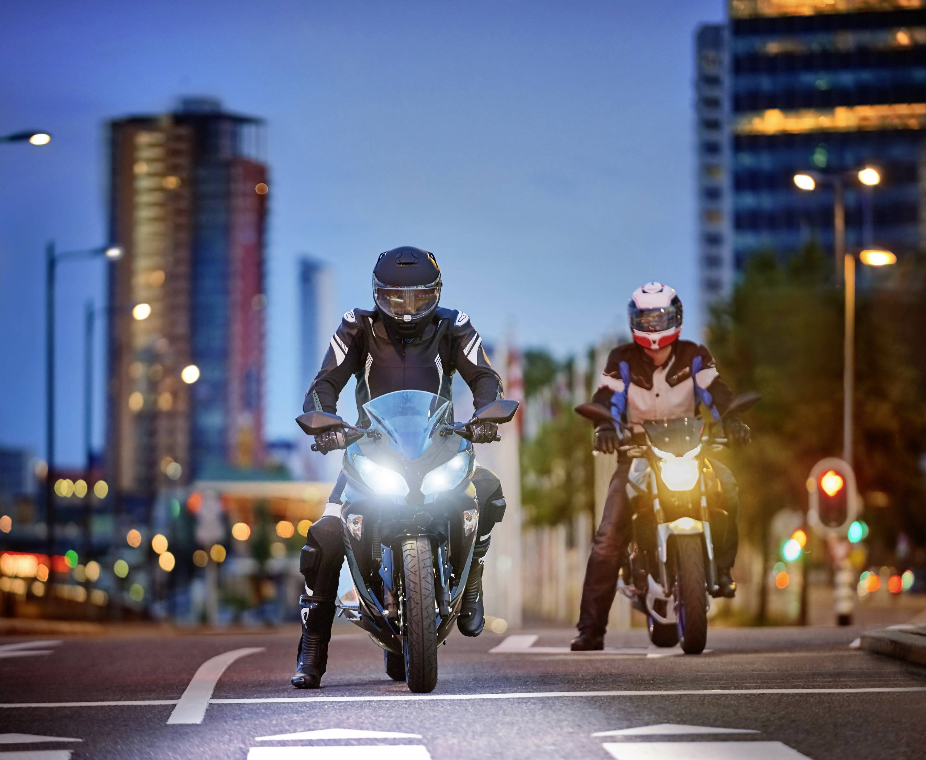 Deux motards roulent sur une route urbaine au crépuscule. Des immeubles illuminés sont visibles en arrière-plan.