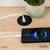 Un smartphone se charge sans fil sur une table en bois à côté d'une plante. Un câble relie la base de charge à une prise électrique.