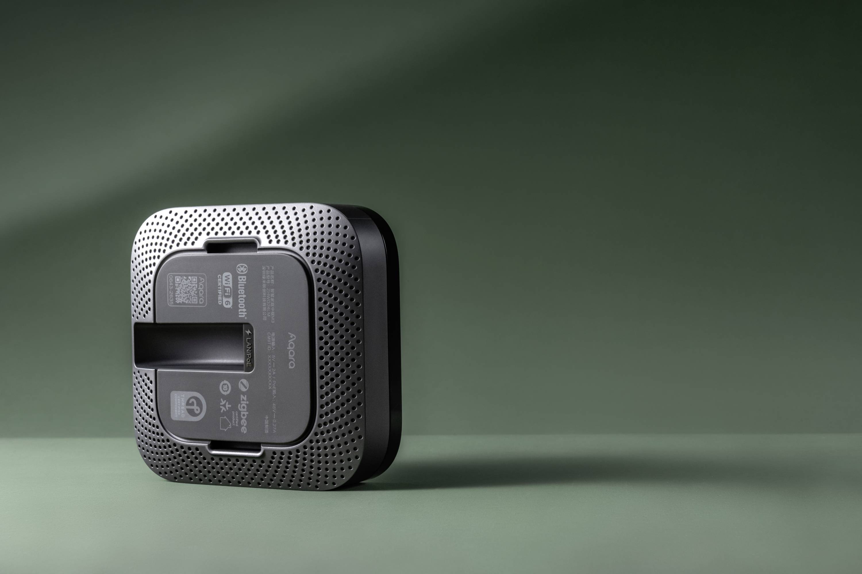 Le dessous d'un haut-parleur carré noir sur un fond vert, avec des ouvertures de ventilation et des informations sur le produit visibles.