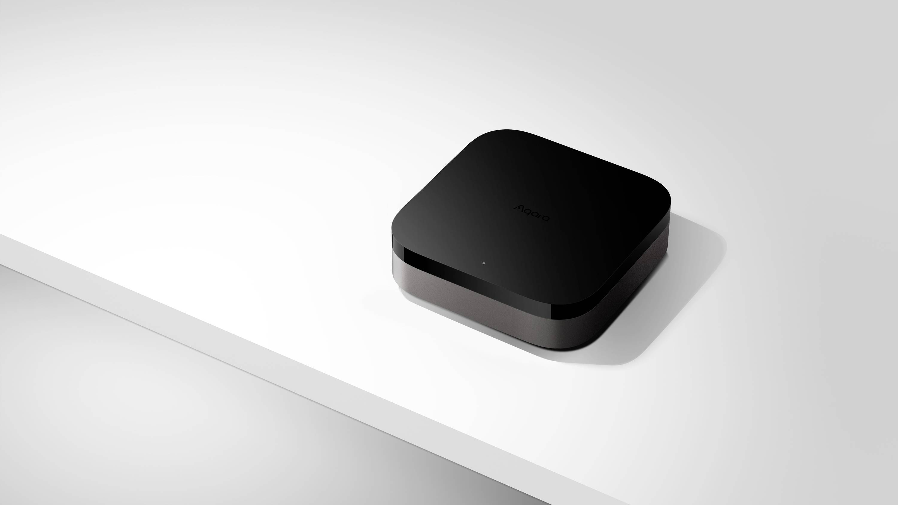 Un appareil noir, carré, aux coins arrondis est posé sur une table blanche. Il présente une surface lisse et un design minimaliste.
