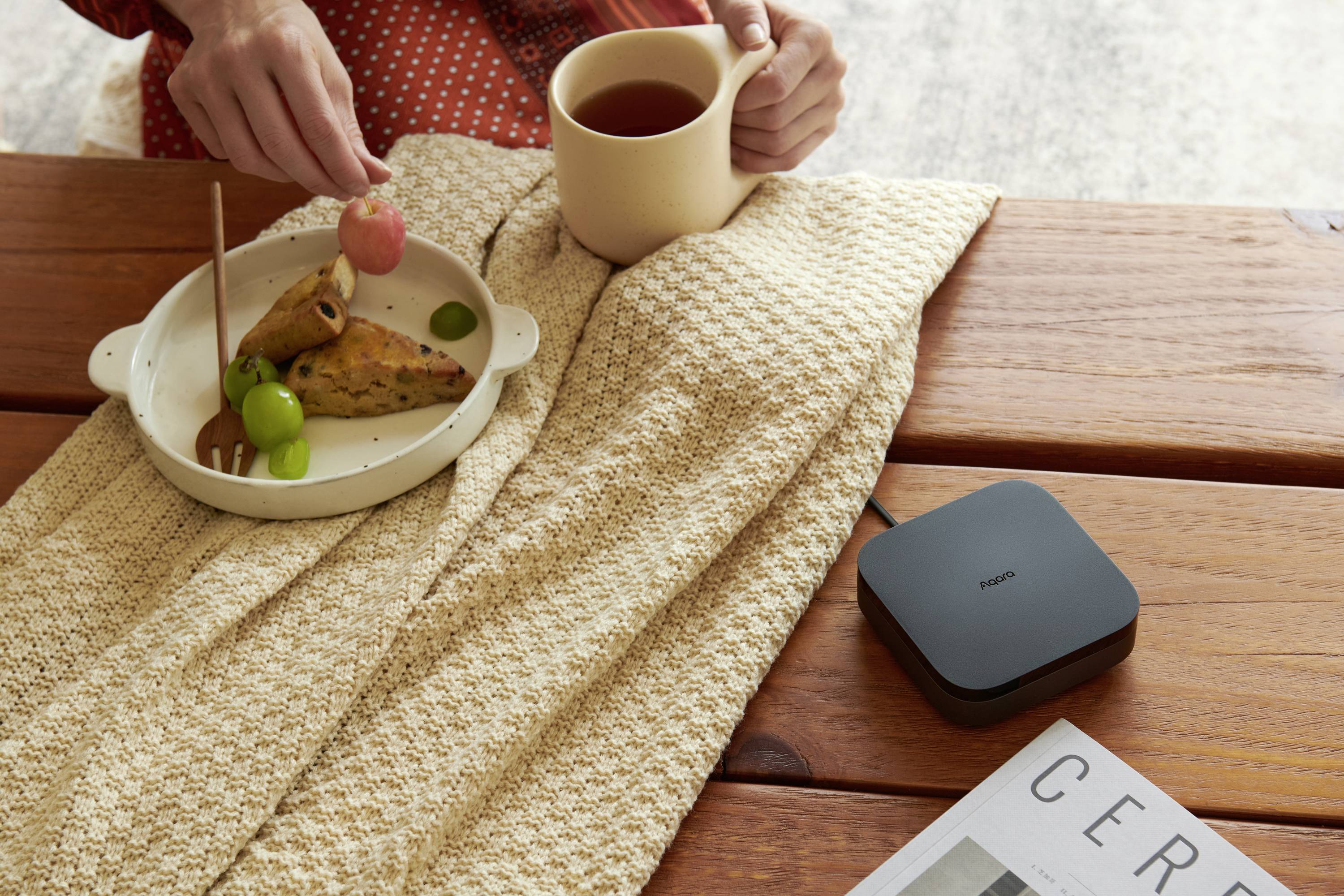 Une personne déguste du thé accompagné d'un petit repas composé de pâtisseries et de raisins. À côté se trouvent un appareil électronique et un magazine.