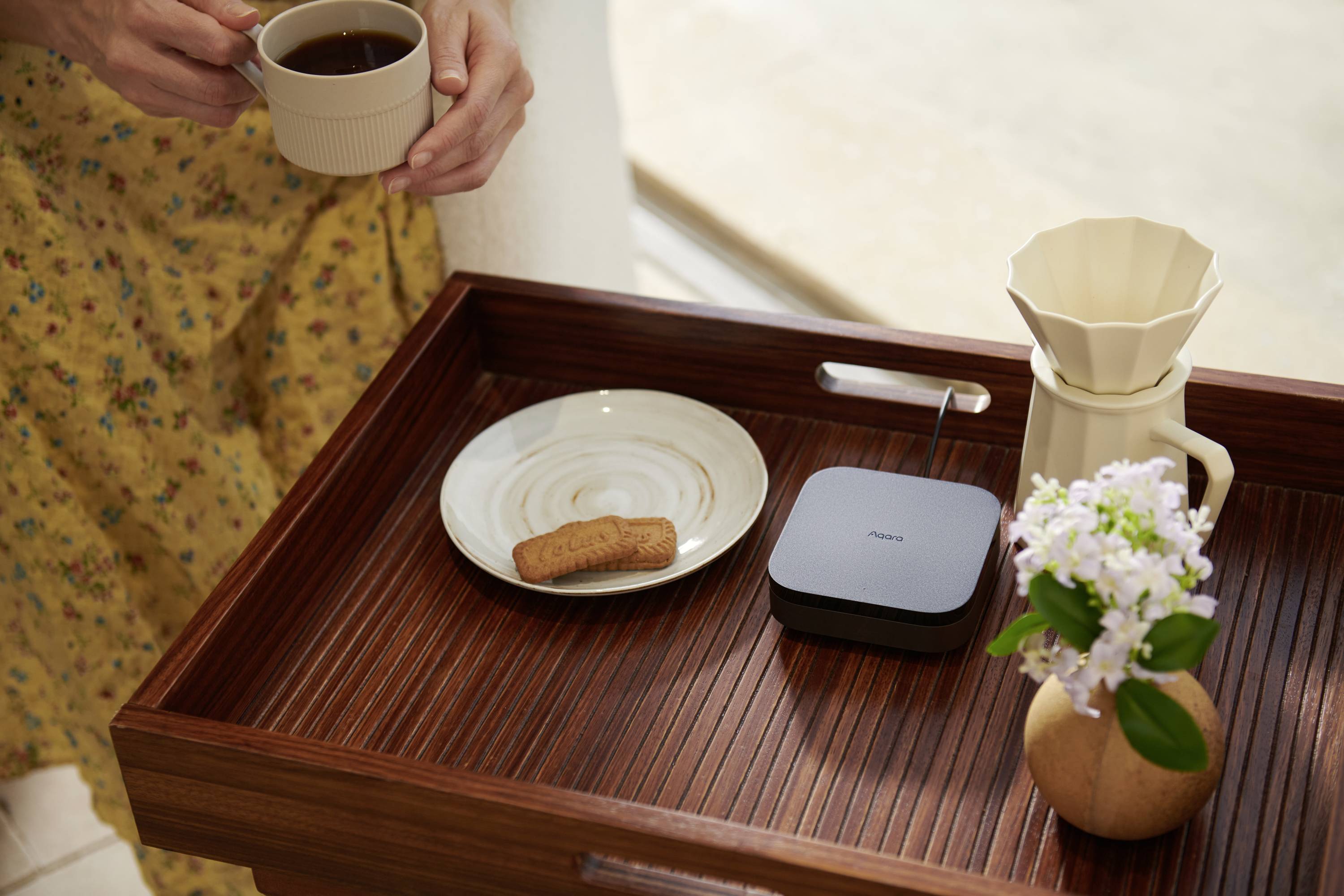 Une personne tient une tasse de café au-dessus d'un plateau comprenant un biscuit dans une assiette, une carafe et un petit appareil.