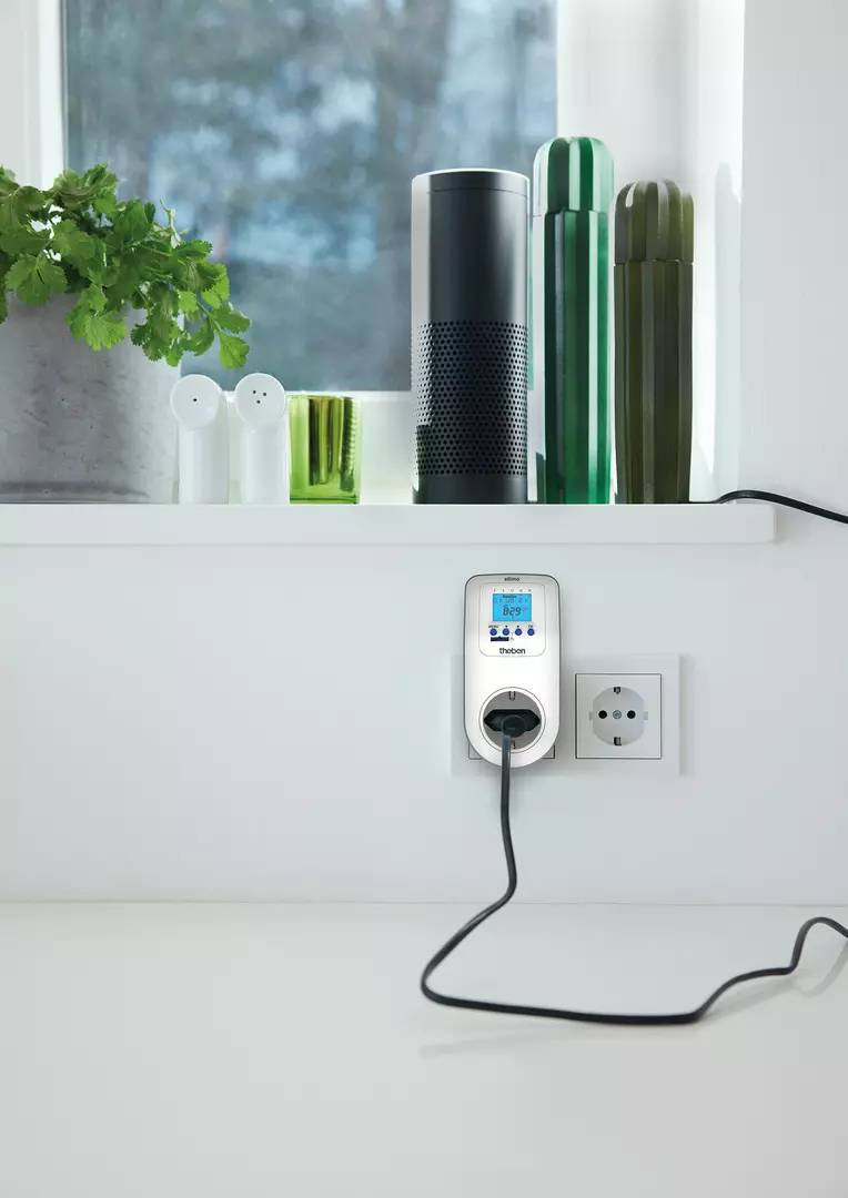 Un compteur intelligent est branché à une prise électrique et affiche la consommation d'énergie. En arrière-plan, des plantes sont posées sur le rebord de la fenêtre, à côté d'un haut-parleur.